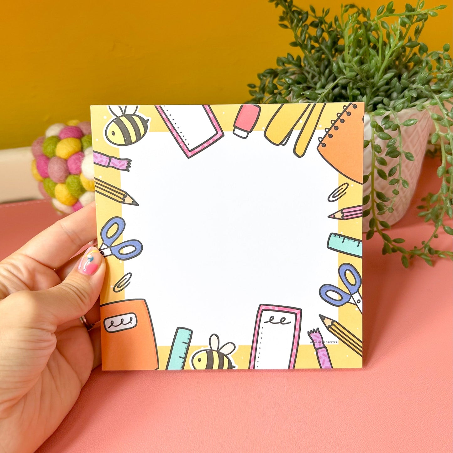 Hand holding a colorful notepad with stationery illustrations on a pink surface with a plant in the background.