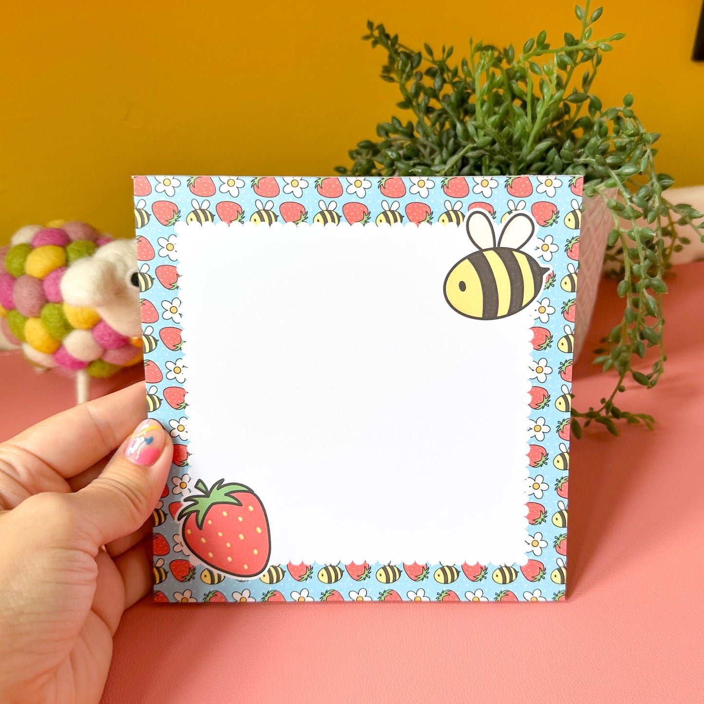 Small square notepad with a colorful design of bees and strawberries held by a hand against a pink and yellow background.