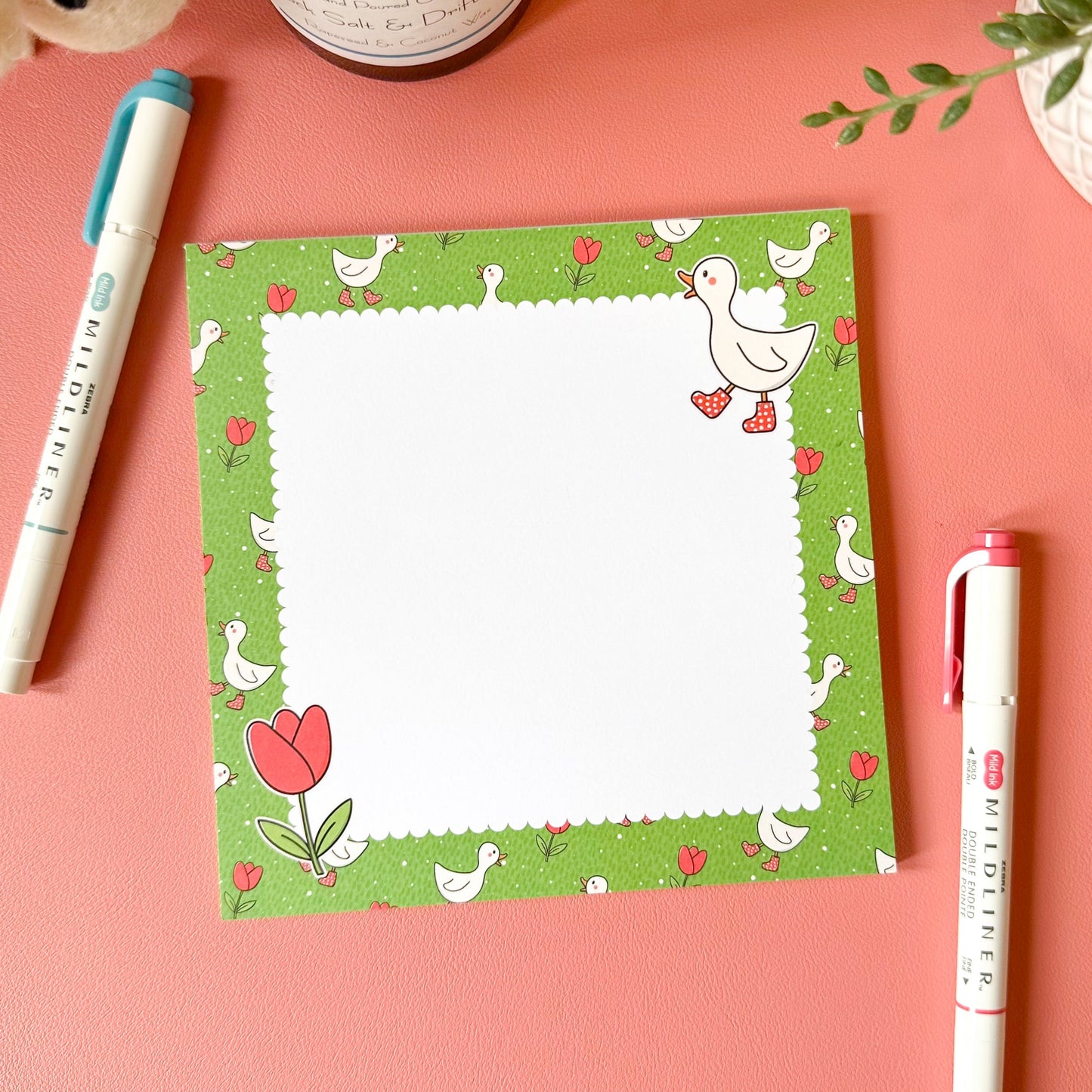 Notepad with a floral design on a pink surface with pens and a candle.