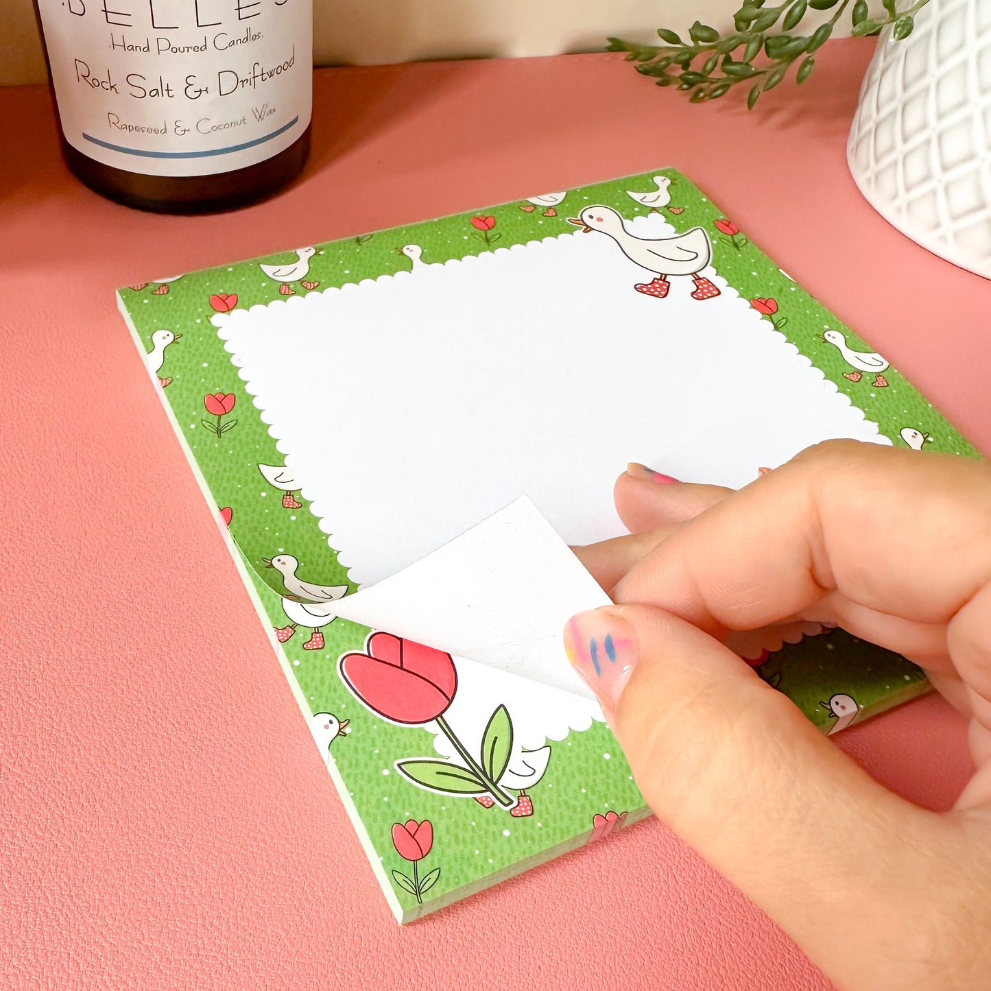 Hand holding a card with a tulip design on a pink surface next to a Belles candle.