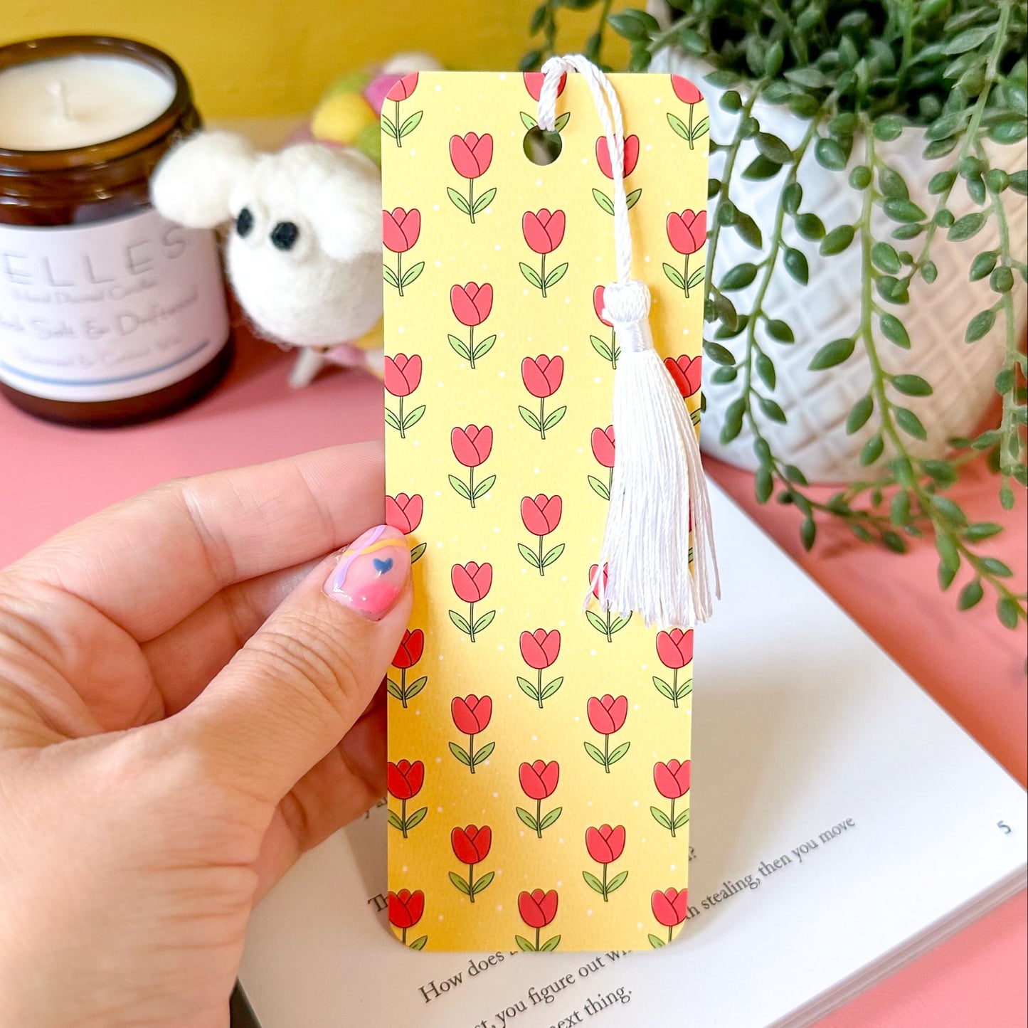 Hand holding a yellow bookmark with red tulip pattern on a book with a yellow background and plants.
