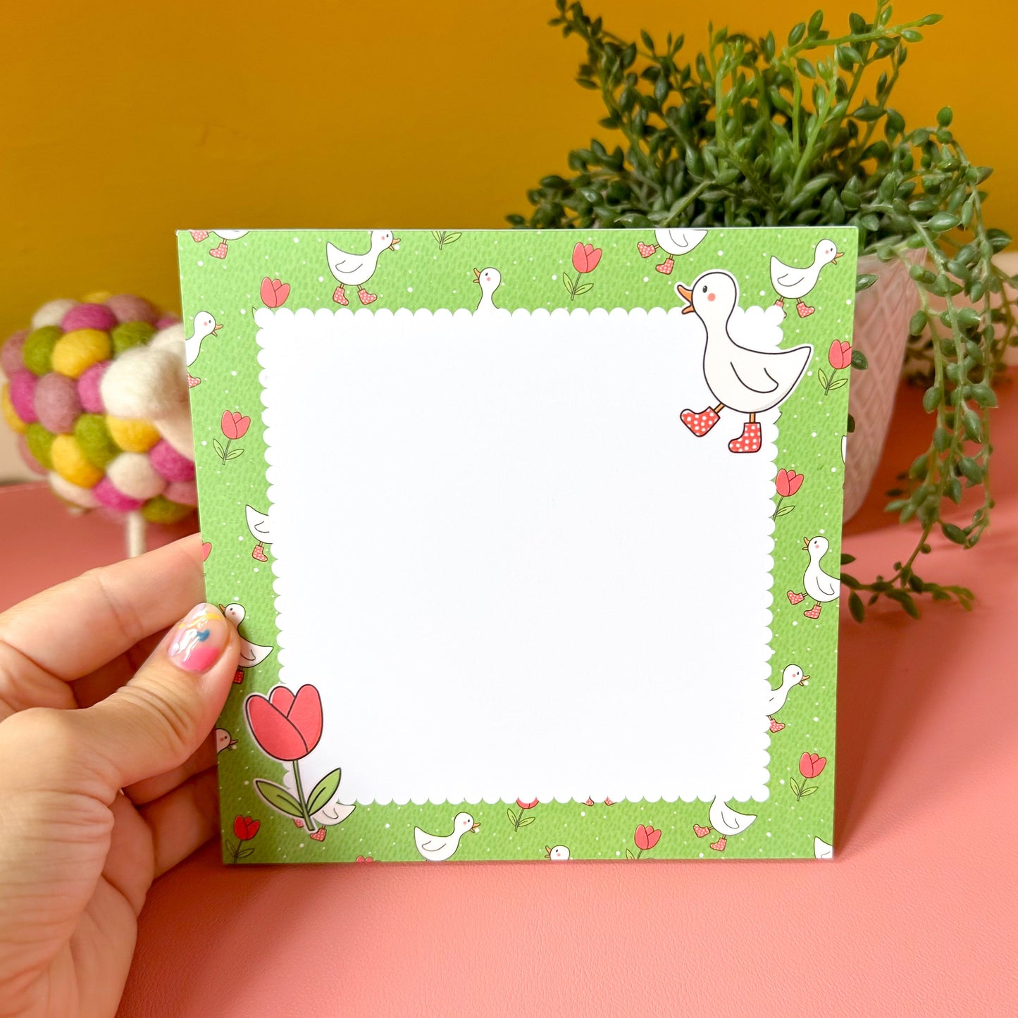 Hand holding a green notepad with duck and tulip designs on a pink surface with plants in the background.