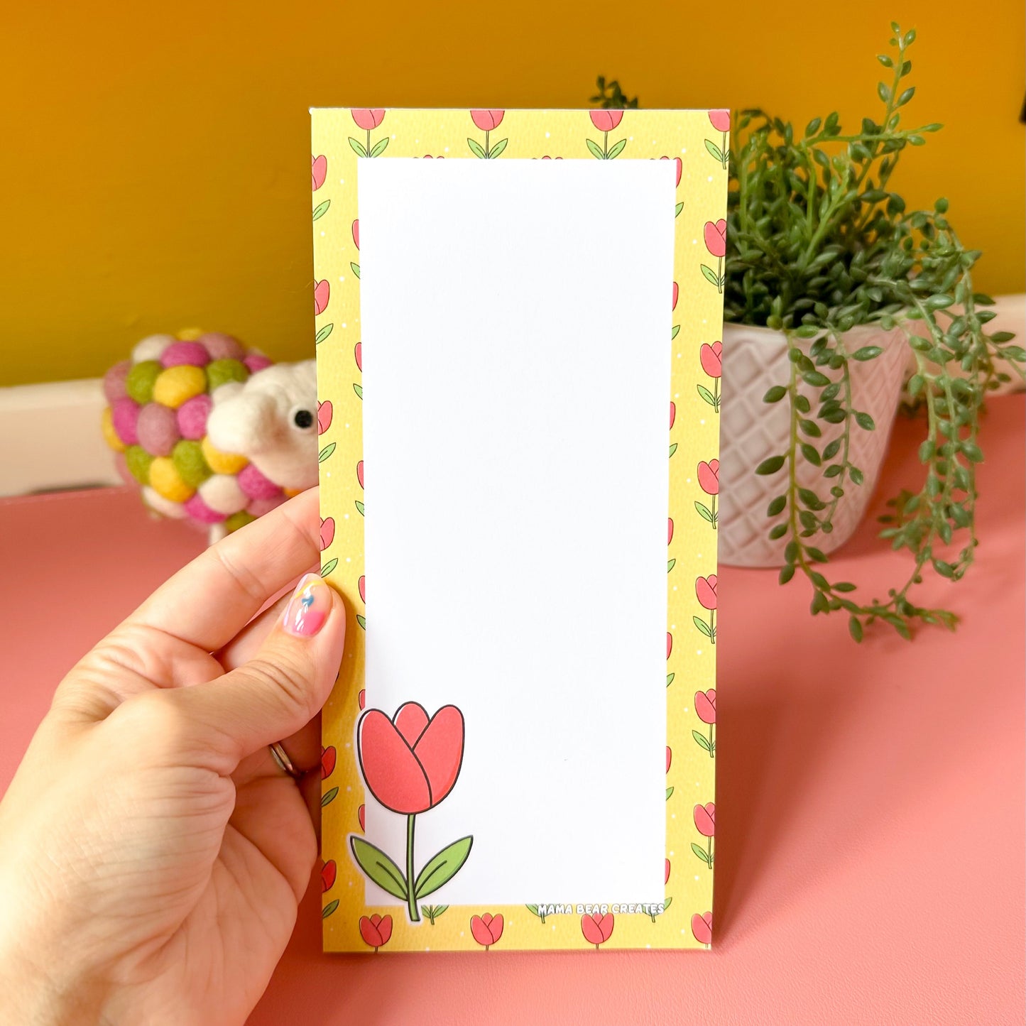 Hand holding a list pad with a tulip design against a yellow and pink background.