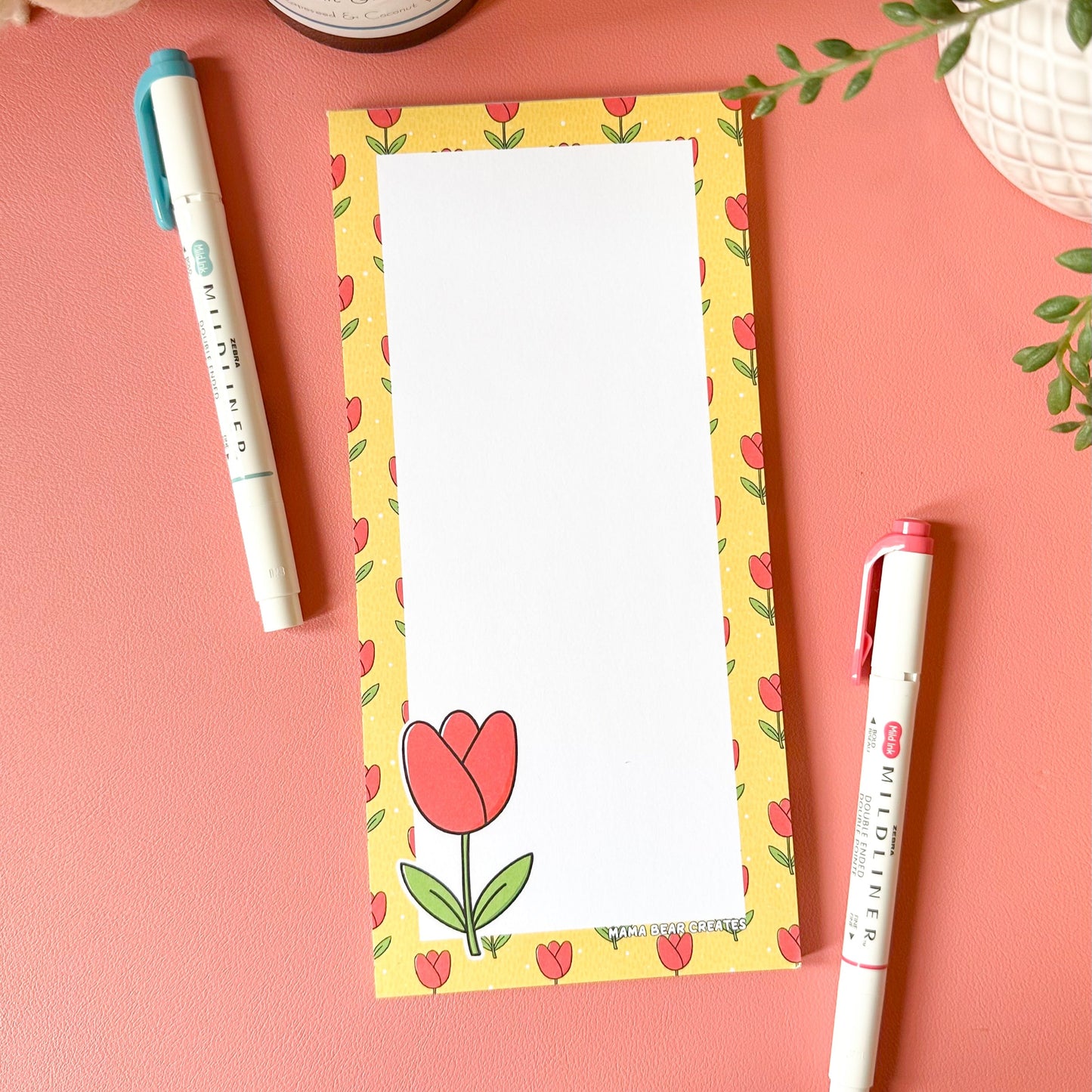 Notepad with a floral design on a pink surface with pens and plants.