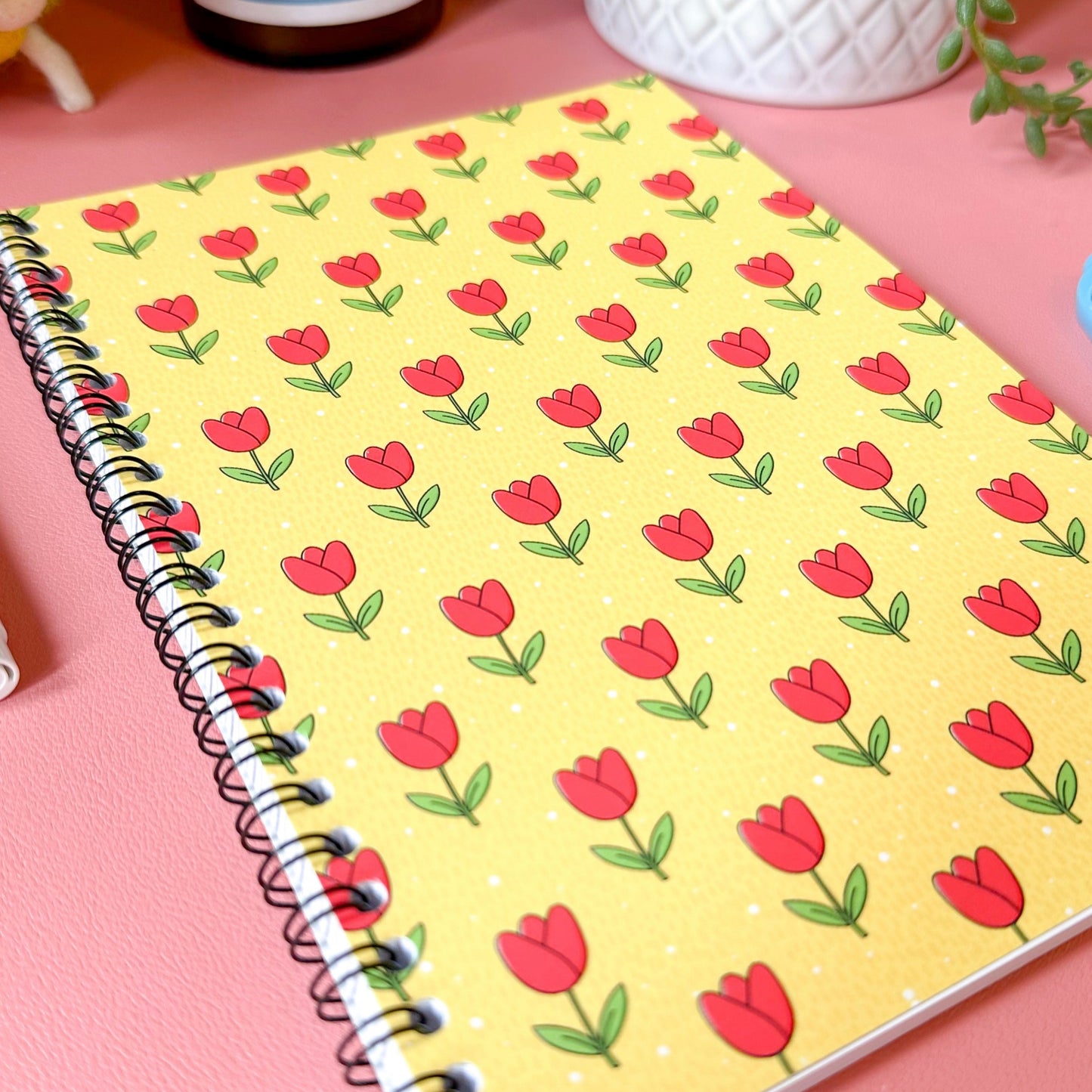Notebook with red tulip pattern on a pink surface with a bottle and plant in the background