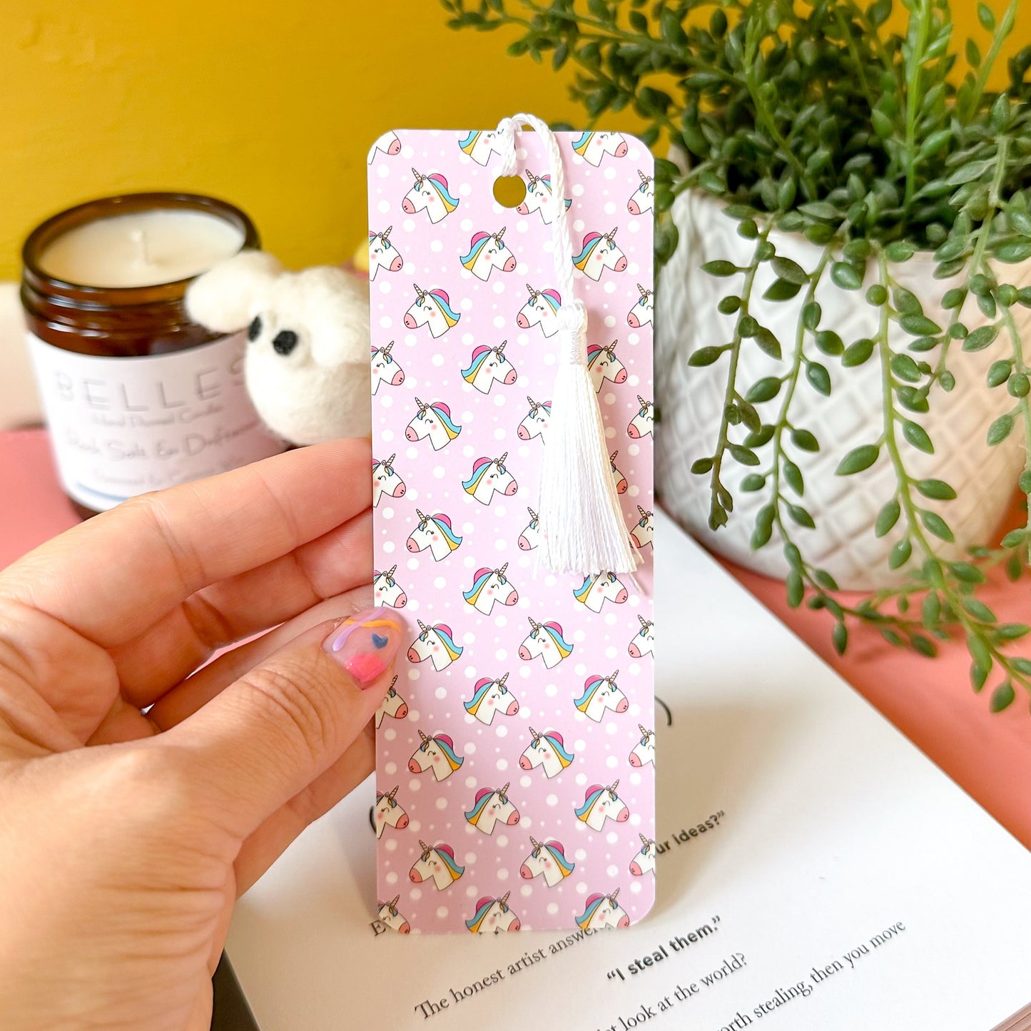 Hand holding a pink bookmark with unicorn pattern on a book with a yellow background