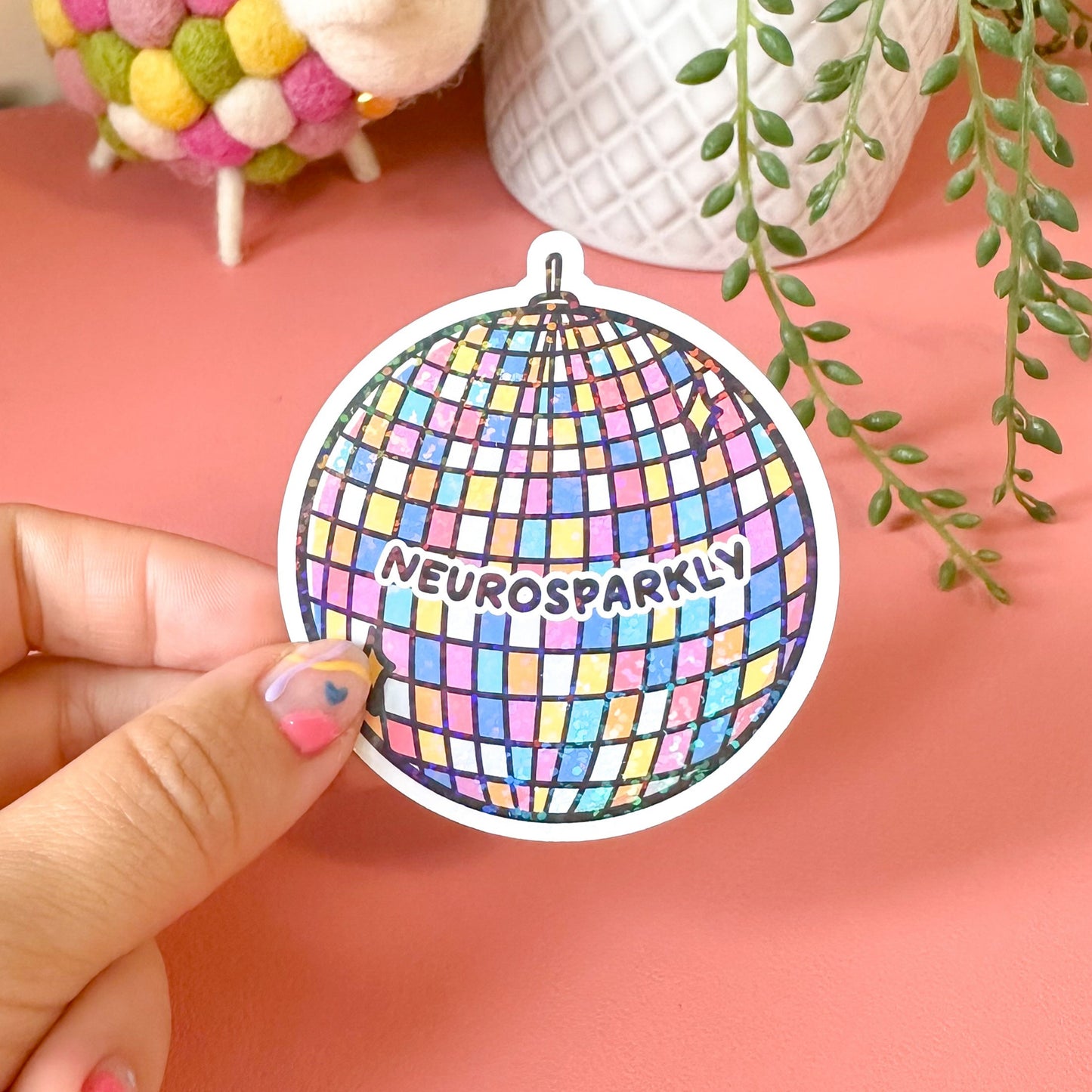 Colorful disco ball sticker with 'Neurosparkly' text held by a hand on a pink surface with decorative items.