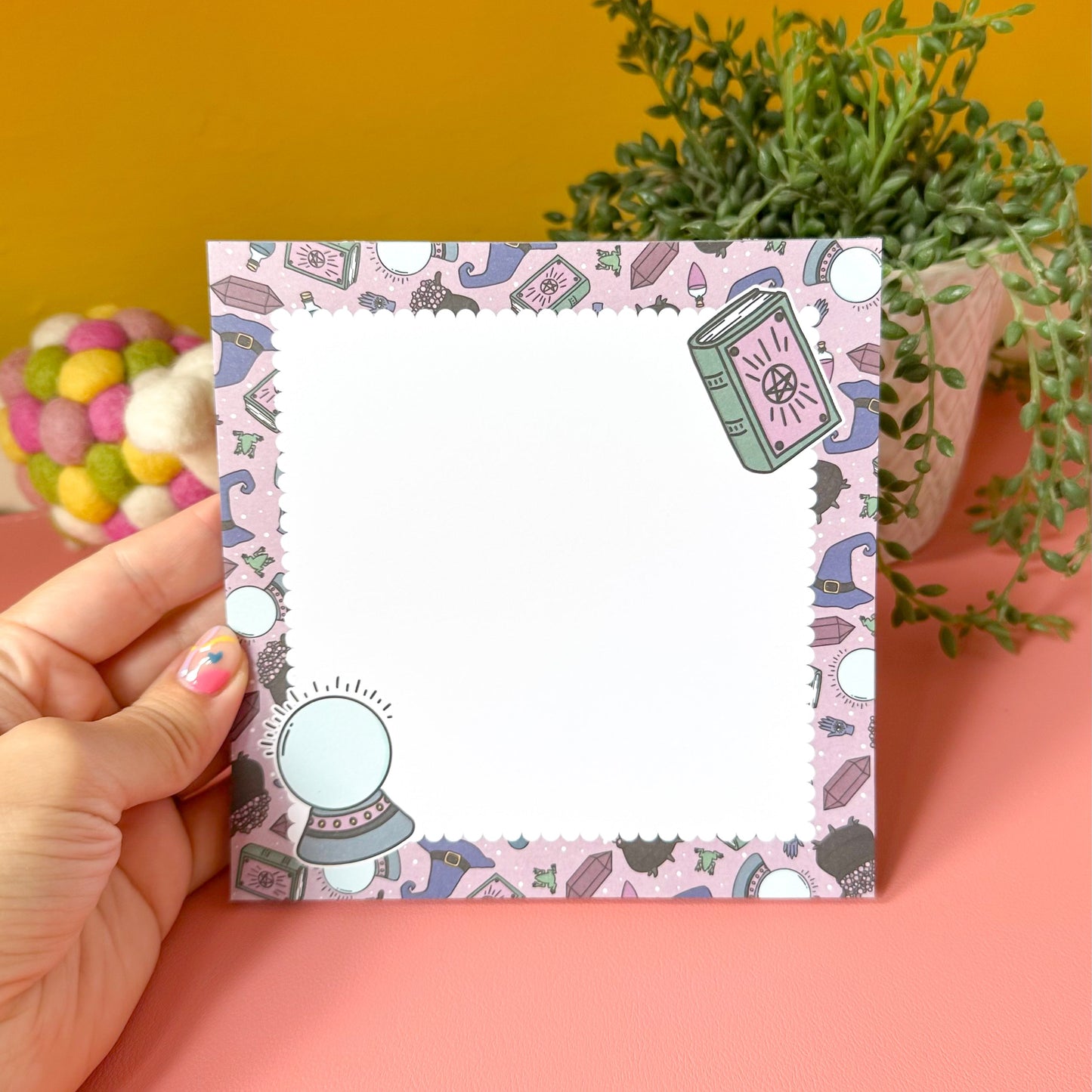Hand holding a small square notepad with a colorful witchy design against a pink background