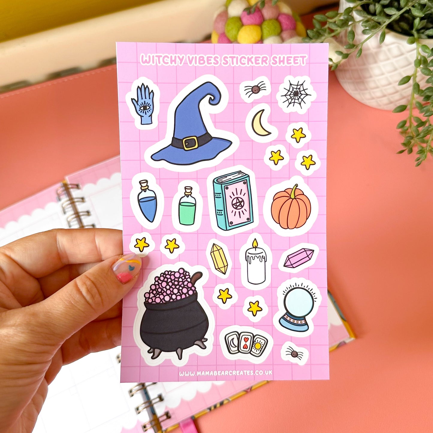Sheet of witch-themed stickers held by a hand on a pink background with a plant and notebook.