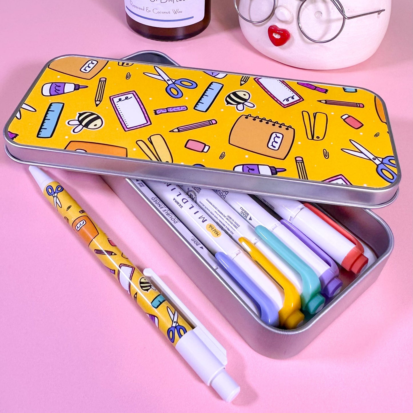 Colorful stationery items in a tin with pens and markers on a pink background