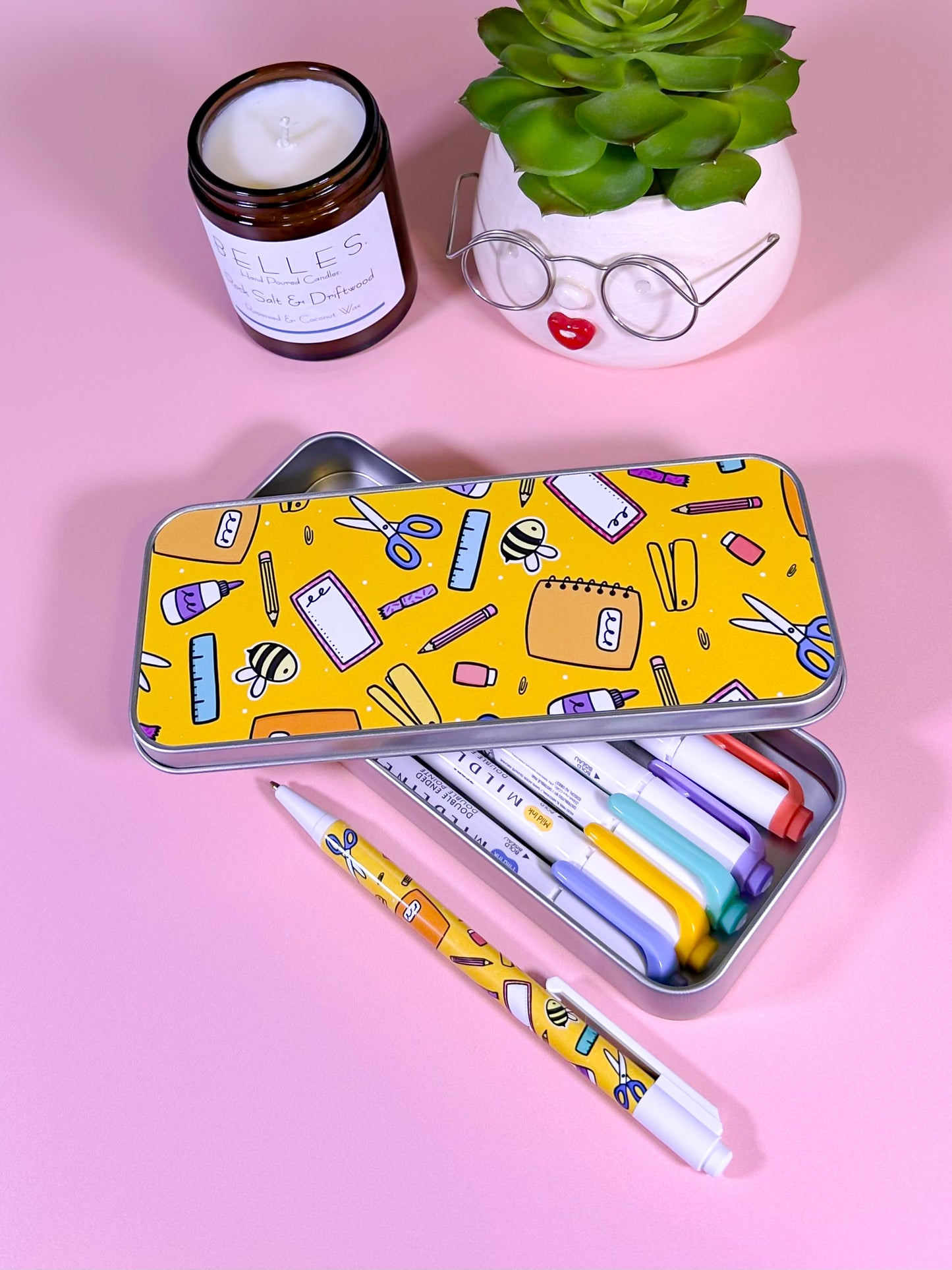 Colorful stationery set with pens and a ruler in a yellow patterned tin on a pink background.