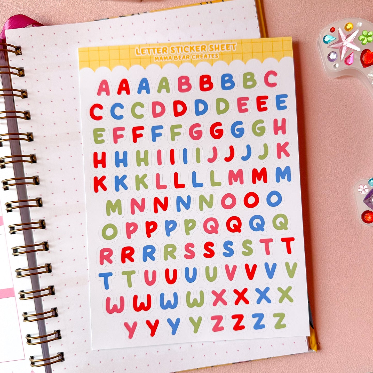 Colorful letter stickers on a spiral-bound notebook with stationery items on a pink background