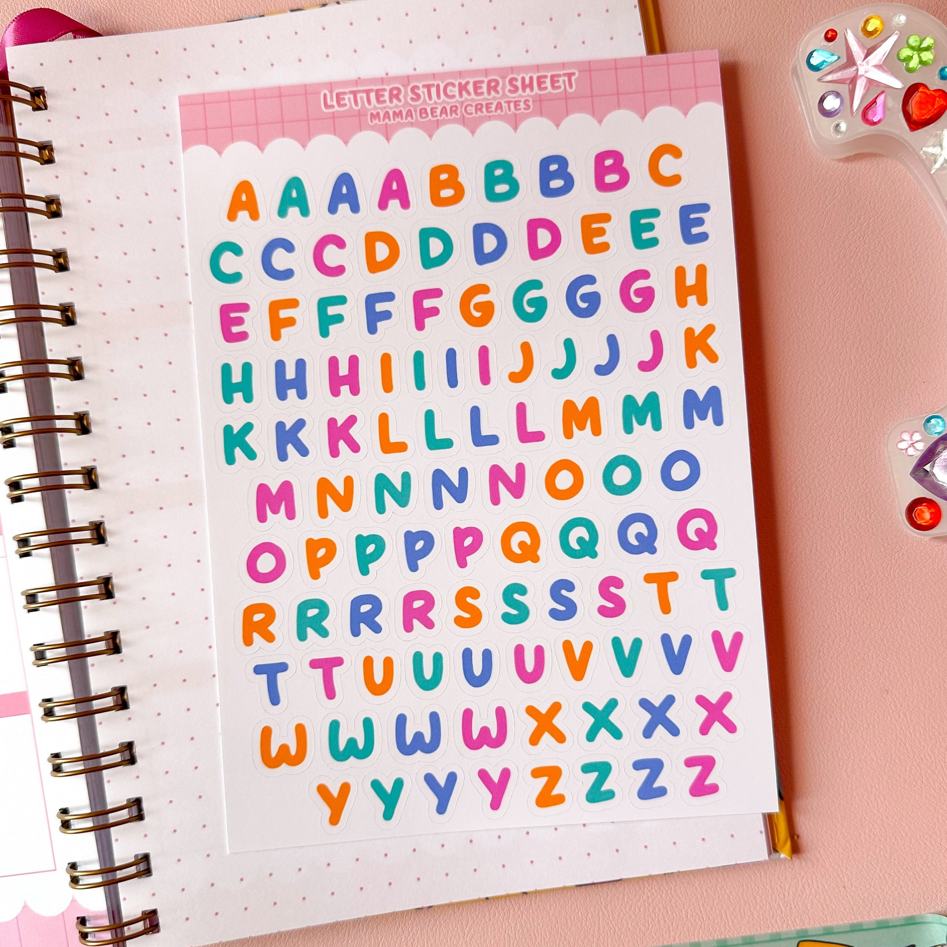 Sheet of colorful letter stickers on a notebook with stationery items on a pink background