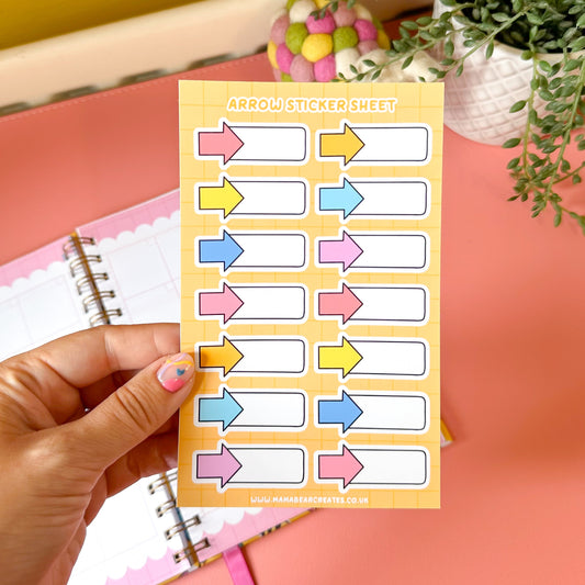 Sheet of colorful arrow stickers held by a hand with a pink notebook and plant in the background.