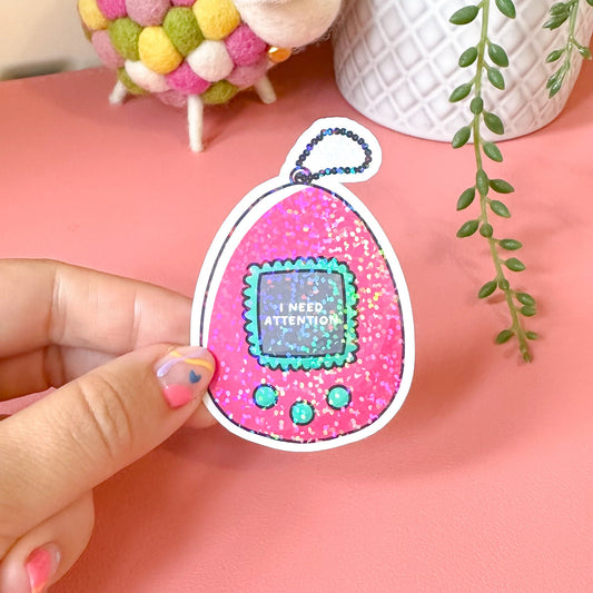 Hand holding a colorful Tamagotchi-style sticker on a pink surface with decorative items in the background.