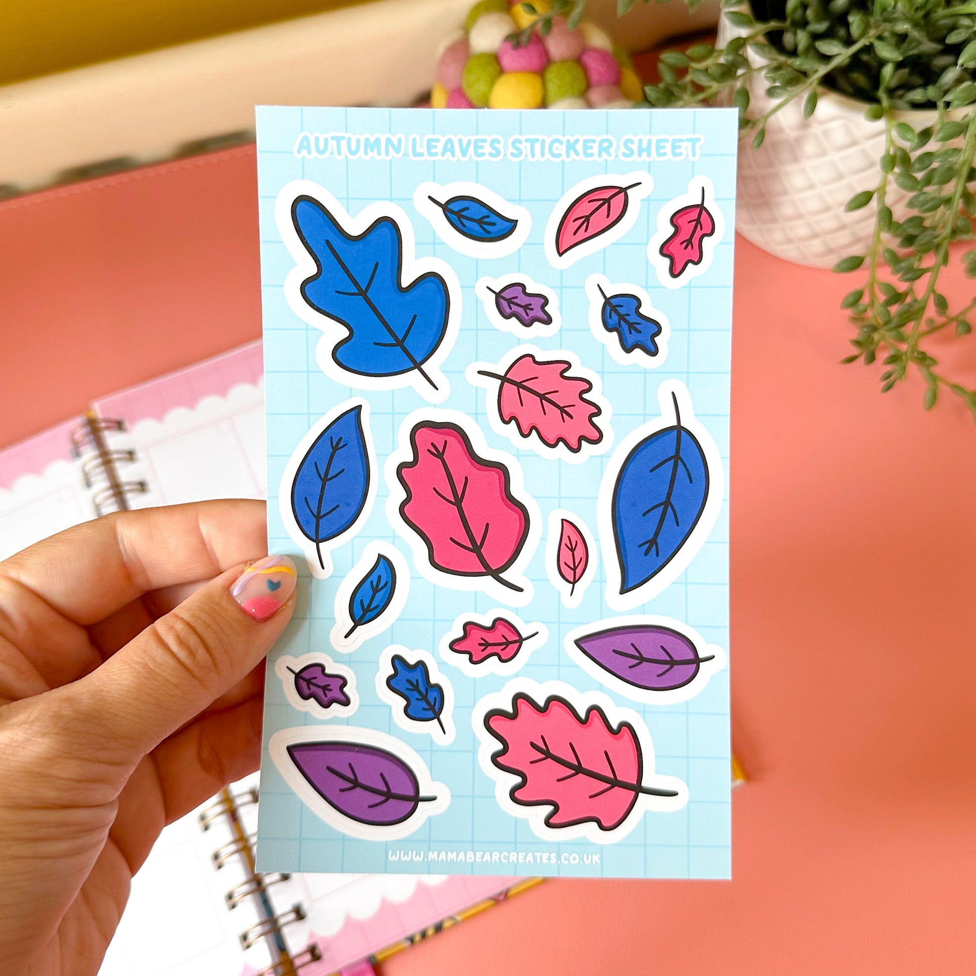 Sheet of colorful autumn leaf stickers held by a hand with a notebook and plant in the background.
