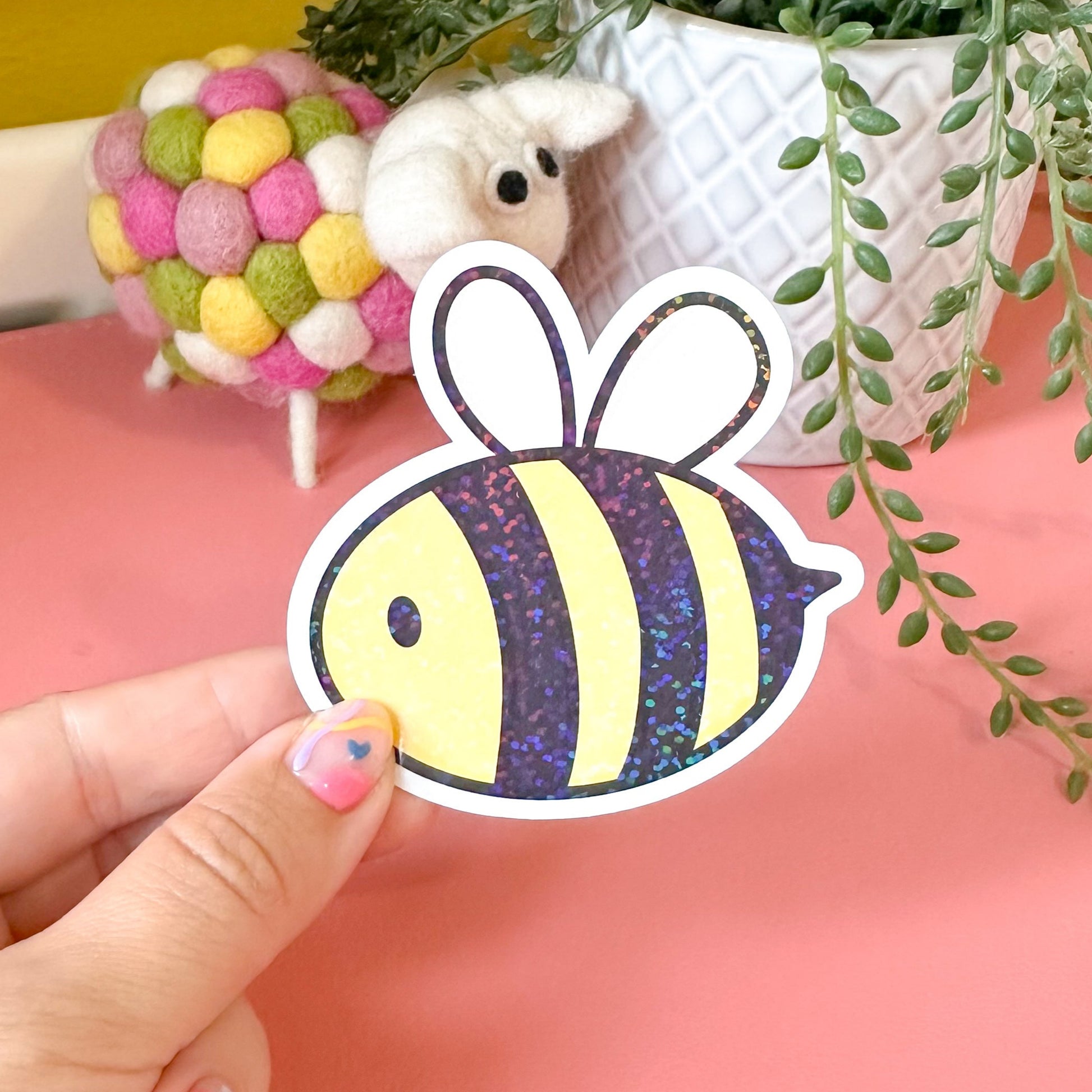 Hand holding a colorful bee-shaped sticker with a pink background and decorative items.