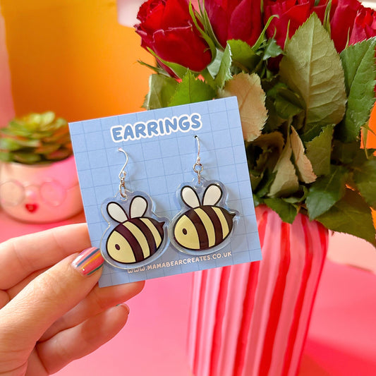 A pair of bee-shaped earrings on a blue earring card, displayed in front of a bouquet of roses.