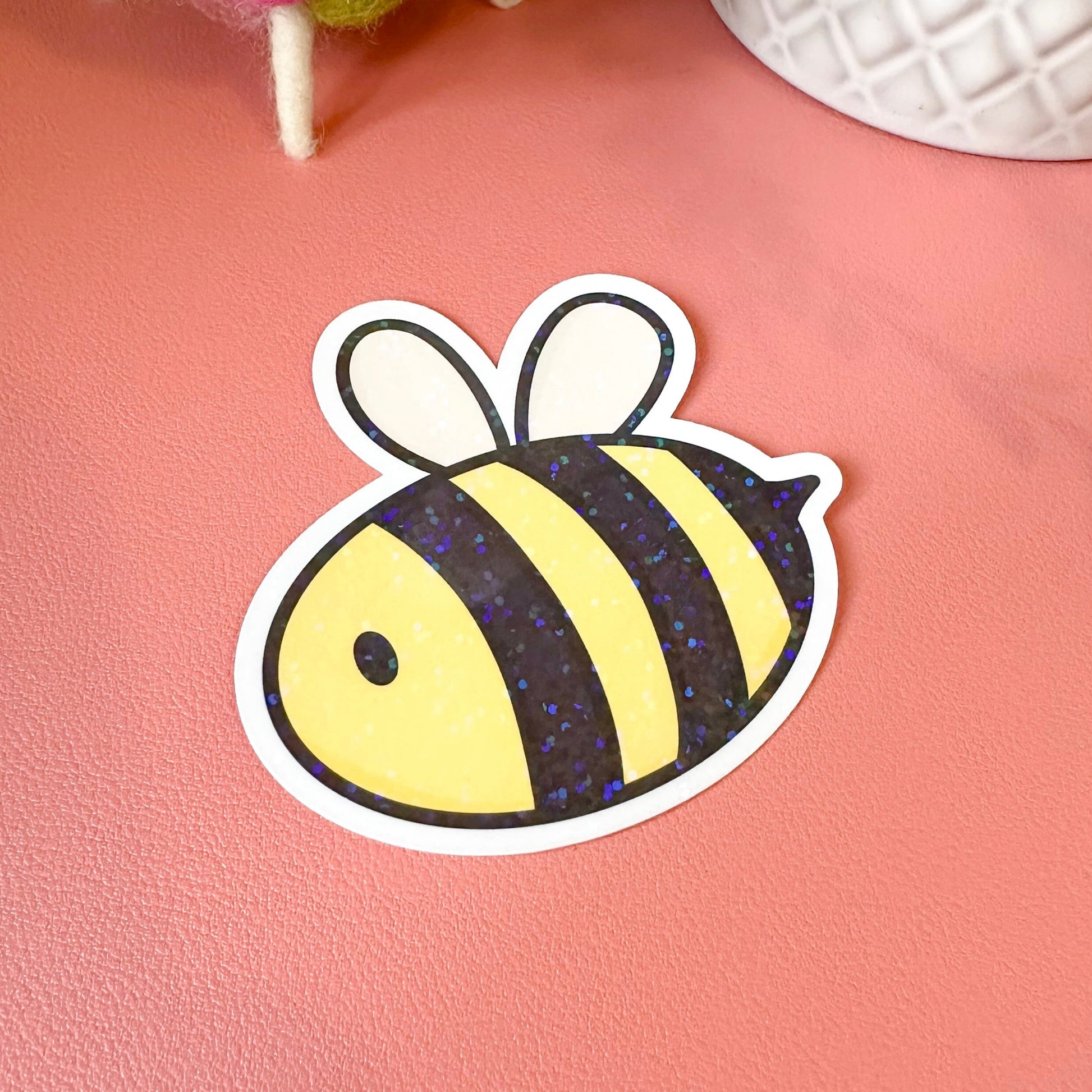 Sticker of a bee on a pink surface with a textured white object and colorful yarn in the background.
