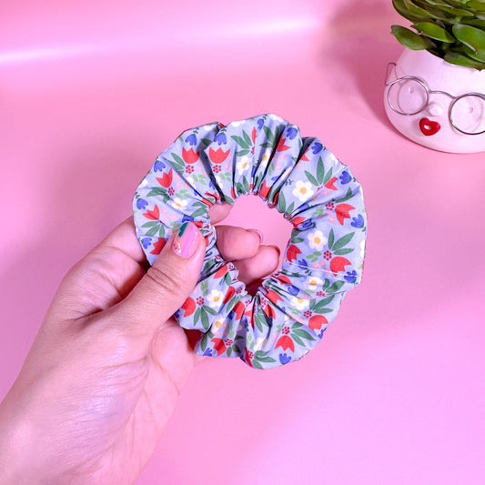 Hand holding a floral scrunchie against a pink background with a small plant and glasses.