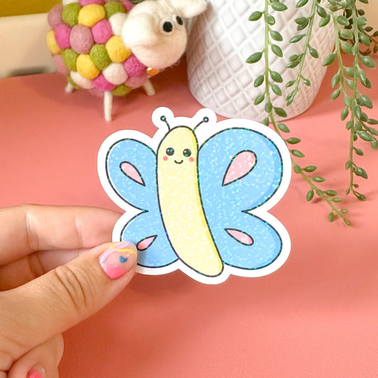 Hand holding a colorful butterfly-shaped sticker with a plant and small toys in the background.