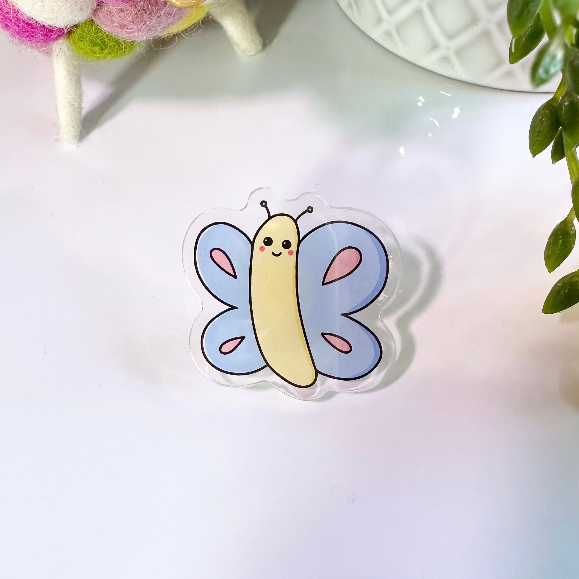 A colorful acrylic pin badge shaped like a butterfly with a smiling face, placed on a flat surface.