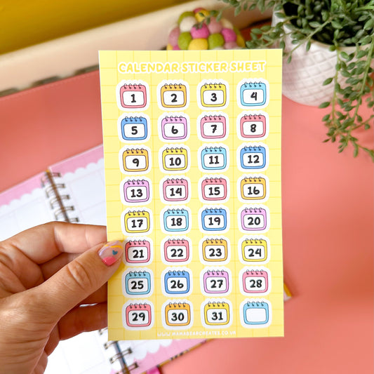 Sheet of colorful calendar stickers held by a hand with a pink notebook and plant in the background.