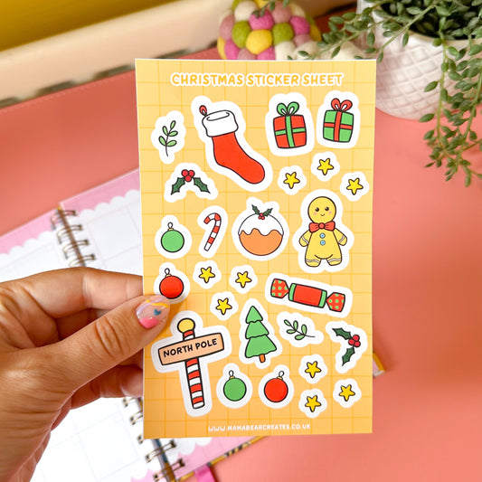Sheet of Christmas-themed stickers held by a hand with a pink notebook and green plant in the background.