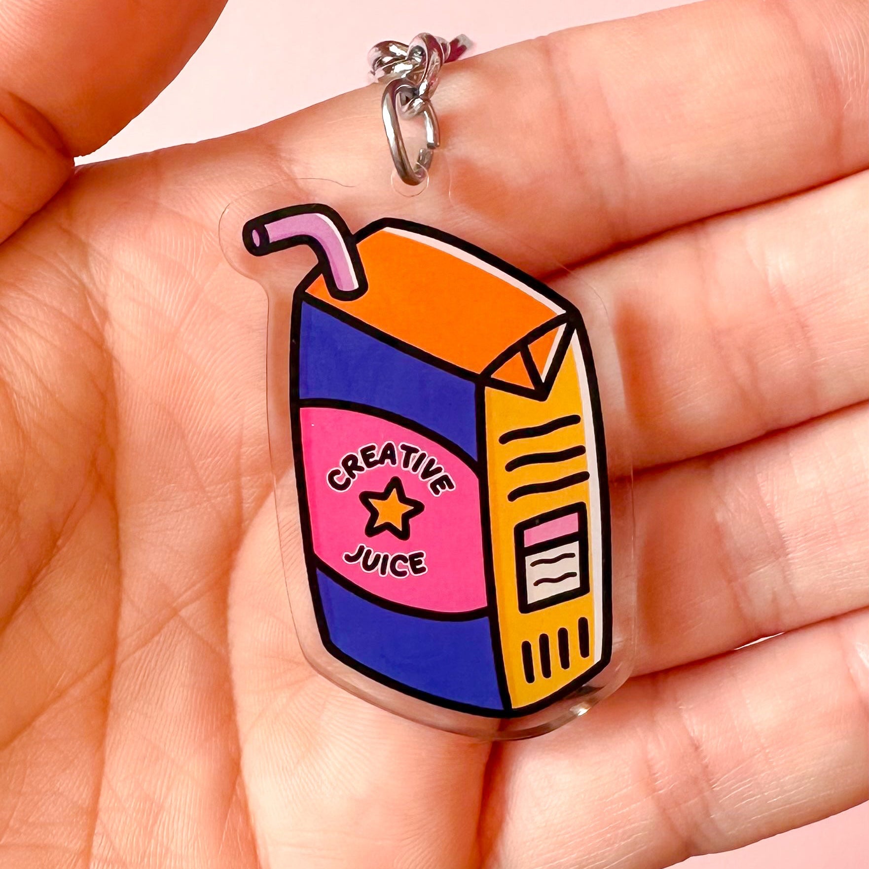 Cartoon-style juice box keychain with 'Creative Juice' text held in a hand against a pink background