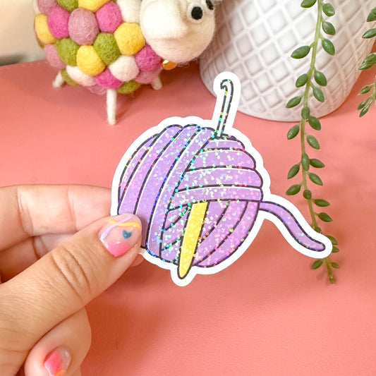 Hand holding a colorful yarn ball sticker with a small white mouse and decorative items in the background.