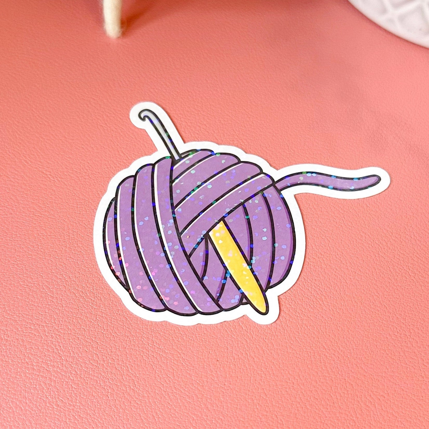 Sticker of a purple yarn ball with a knitting needle on a pink background