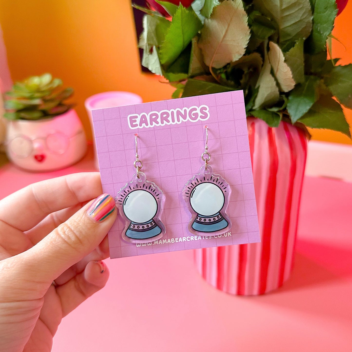 A pair of crystal ball earrings on a pink earring card, with the word 'EARRINGS' printed at the top. The earrings are held in a hand, and there is a plant and a striped container in the background.
