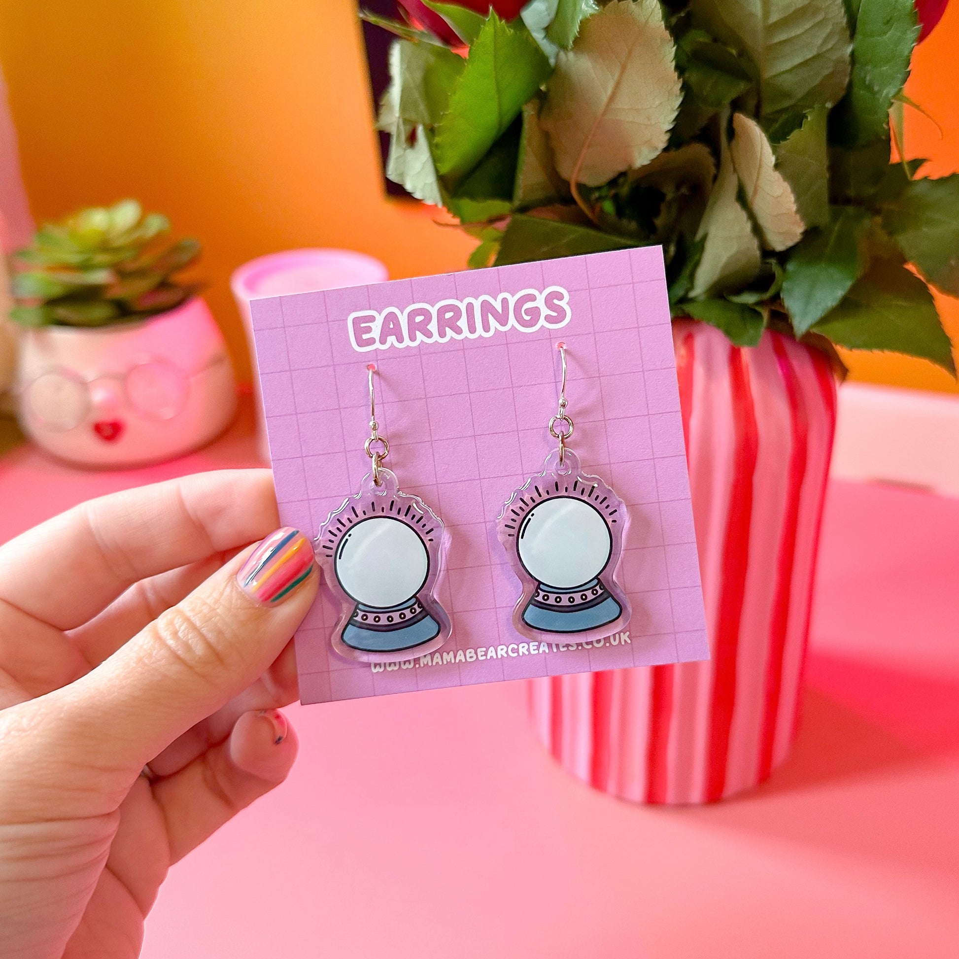A pair of crystal ball earrings on a pink earring card, with the word 'EARRINGS' printed at the top. The earrings are held in a hand, and there is a plant and a striped container in the background.