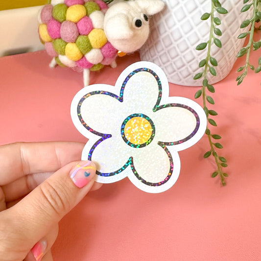 Hand holding a colorful flower-shaped sticker with a small plush toy and plant in the background.