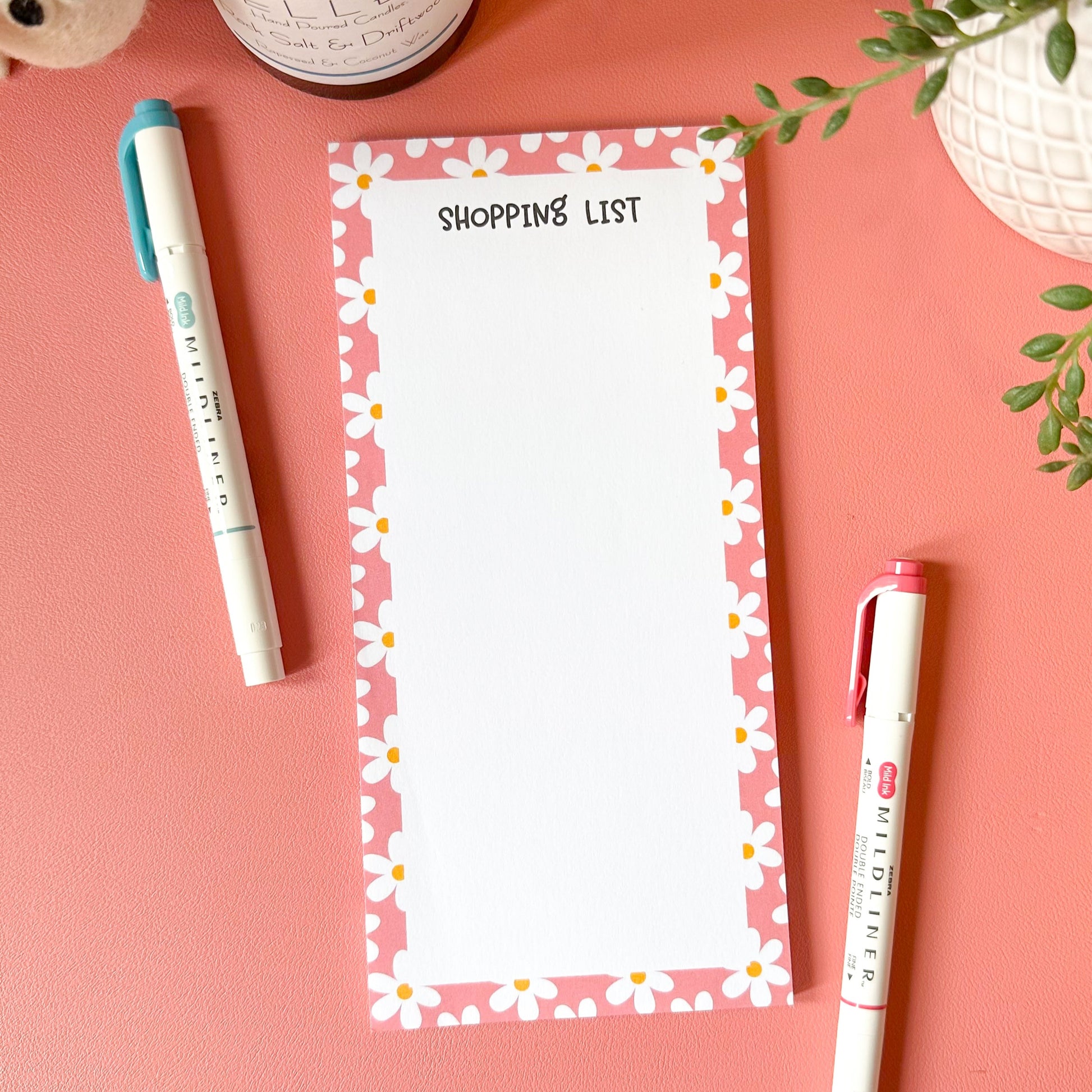 Notepad with 'Shopping List' on a pink surface with pens and a plant.