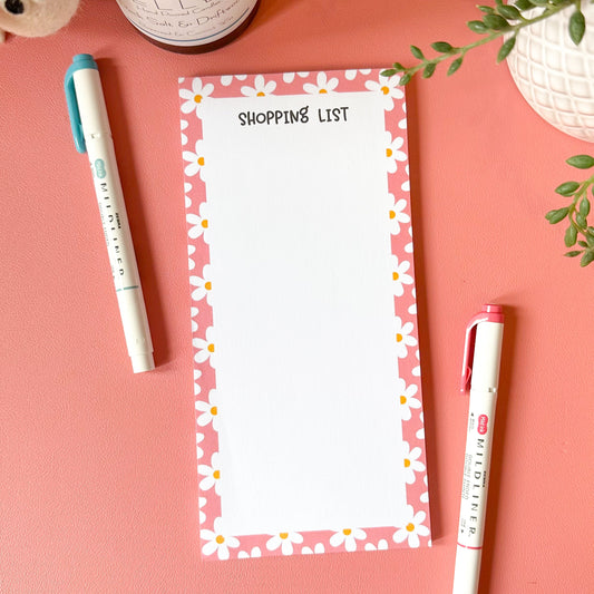 Notepad with 'Shopping List' on a pink surface with pens and a plant.