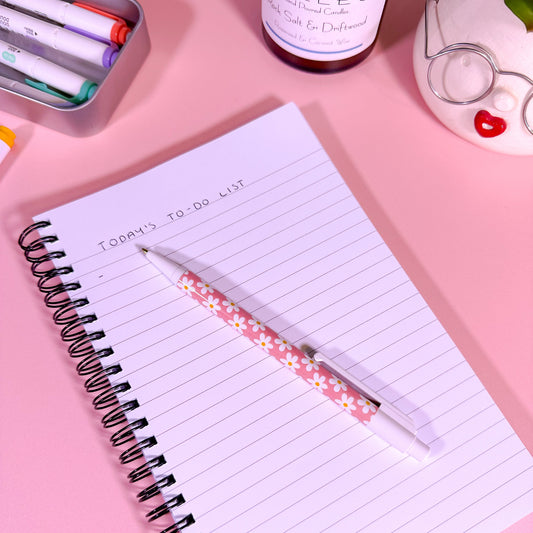 Notebook with a daisy pen on a pink surface with stationery items around