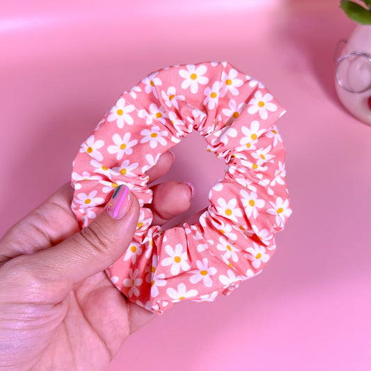 Pink scrunchie with white floral pattern held by a hand against a pink background