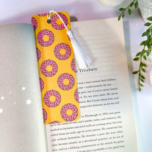 A yellow bookmark with pink donut patterns is inserted in an open book. It features rounded corners, a white tassel, and is double-sided.