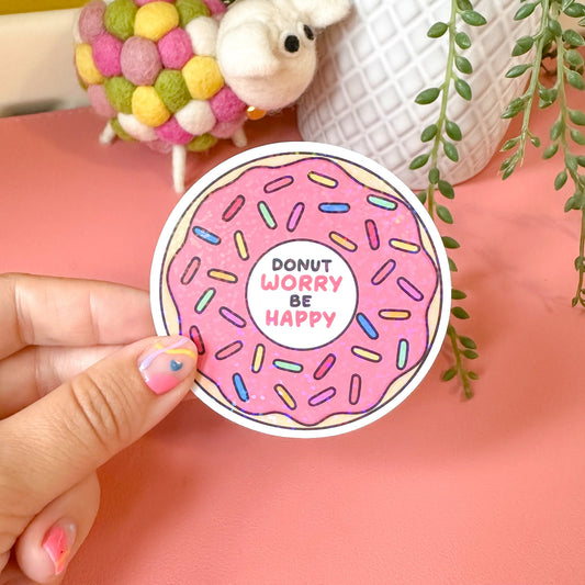 Hand holding a pink donut-shaped sticker with 'Donut Worry Be Happy' text, surrounded by decorative items on a pink surface.