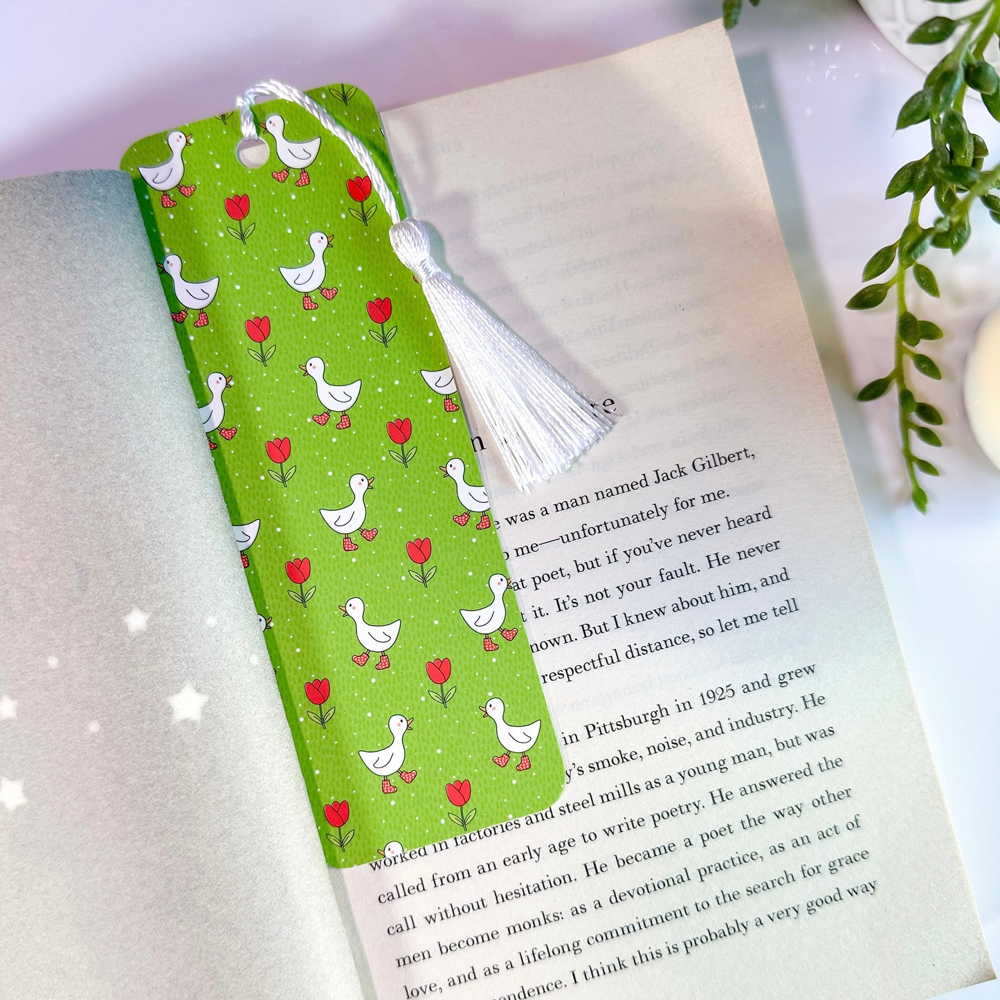 A green bookmark with illustrations of ducks and tulips, featuring a white tassel, is placed within an open book.
