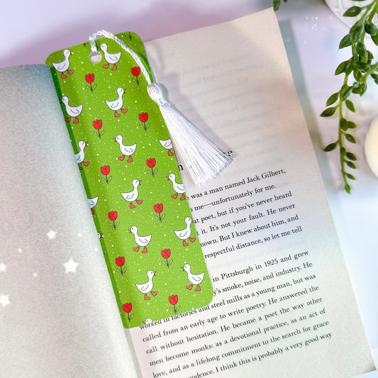 A green bookmark with illustrations of ducks and tulips, featuring a white tassel, is placed within an open book.