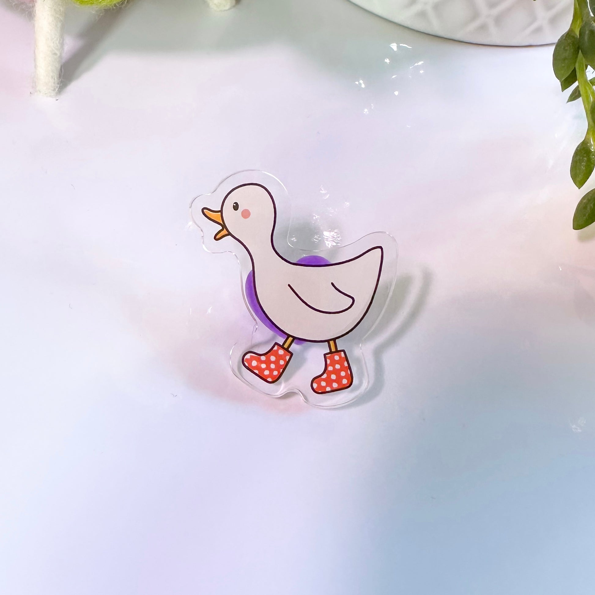 A pin badge featuring an acrylic depiction of a white duck with red boots, presented on a soft background.