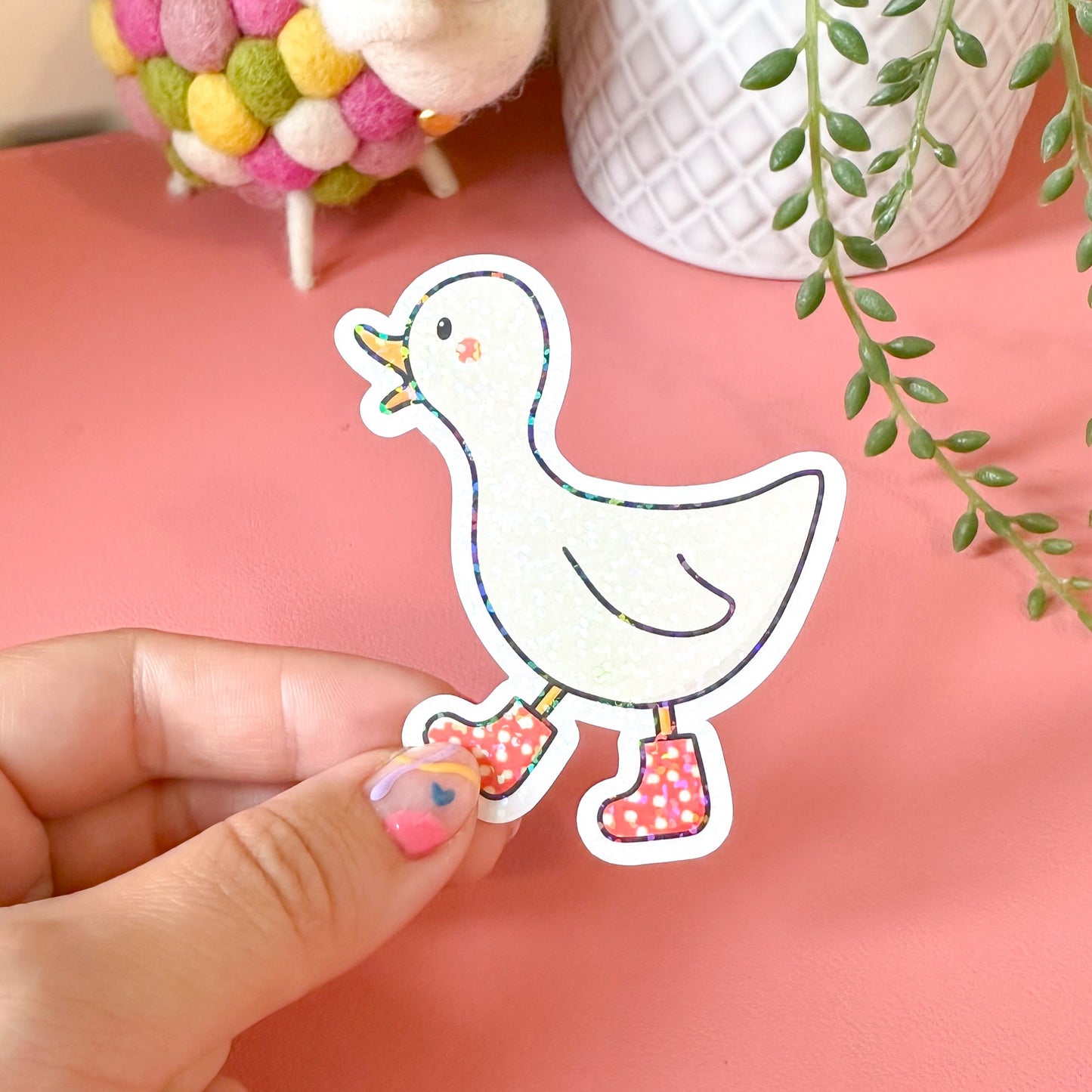 Hand holding a cartoon duck sticker with colorful shoes on a pink surface with plants in the background