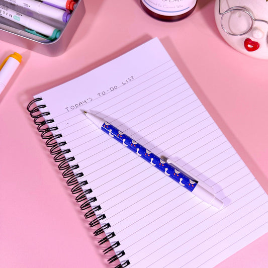 Notebook with a pen on a pink surface with stationery items around