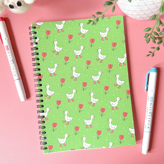 Green notebook with duck and flower pattern on a pink surface with pens and plants.