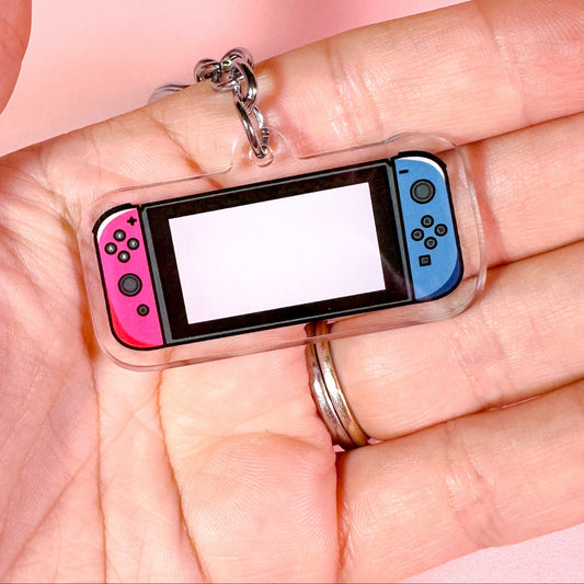 Keychain shaped like a handheld gaming console held in a hand against a pink background