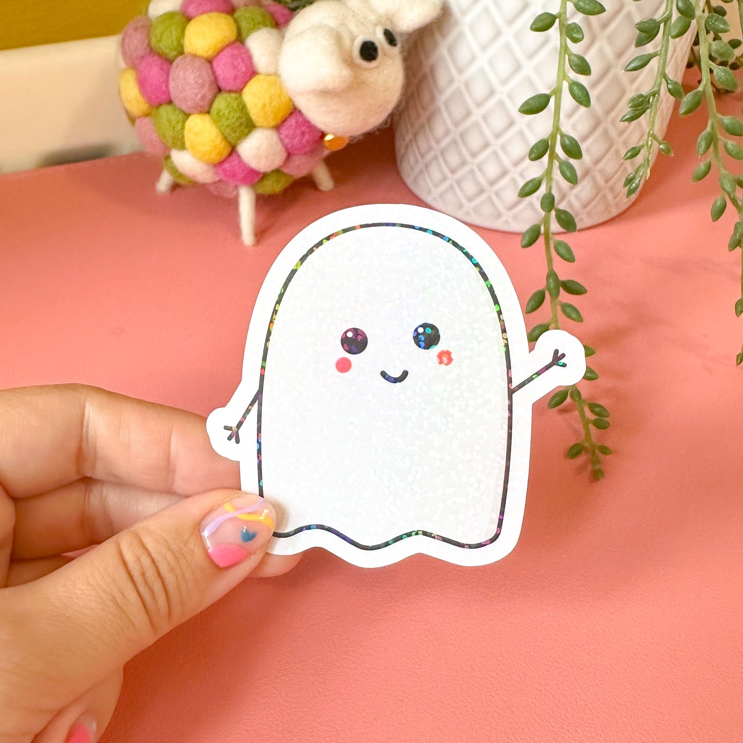 Hand holding a cute ghost sticker with a pink background, plant, and colorful ball.
