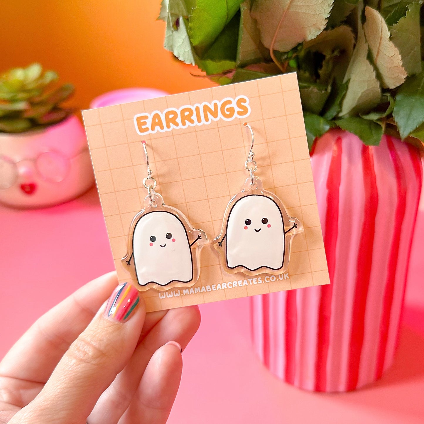 A pair of white acrylic ghost-shaped earrings on an orange earring card, held in a person's hand, with decorative items in the background.