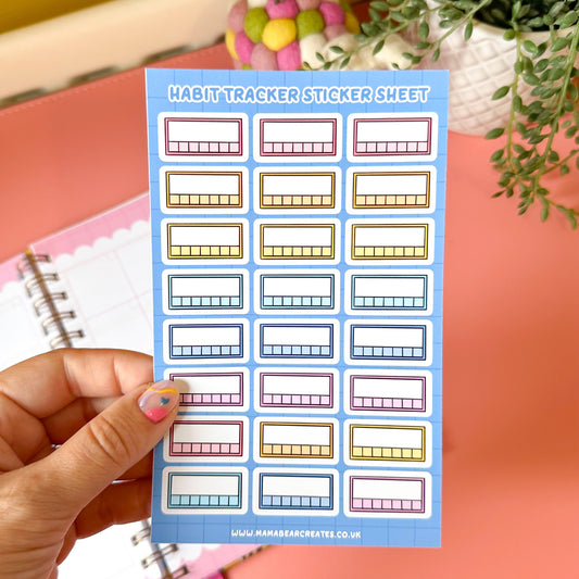 Habit tracker sticker sheet held by a hand with a pink notebook and plant in the background